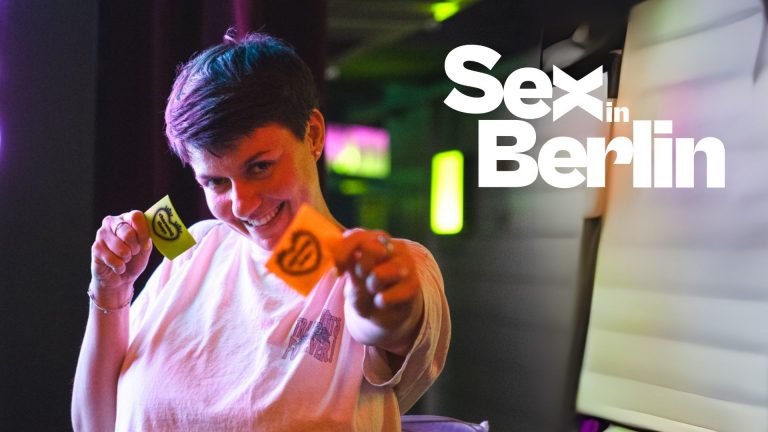 Frau mit kurzem Haar hält zwei farbige Karten in die Kamera, im Hintergrund neonfarbene Lichter und der Titel "Sex in Ber...