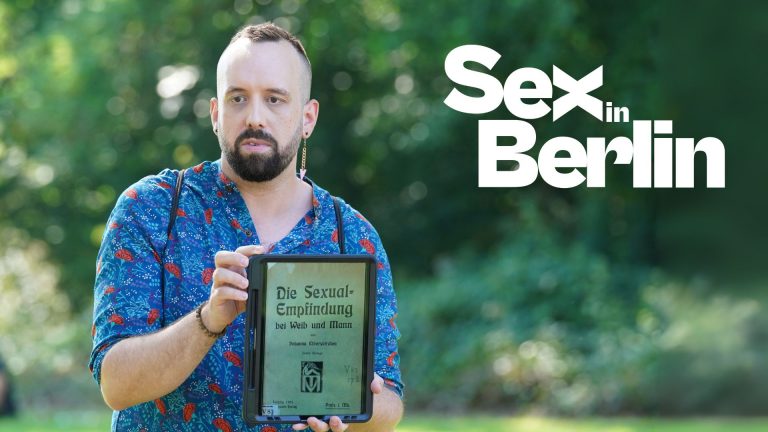 Mann mit Bart hält ein Tablet mit dem Titel "Die Sexual-Empfindung bei Mann und Mann" in einer grünen Parklandschaft.