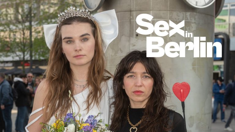 Zwei Frauen stehen vor einer Uhr in Berlin, eine trägt ein Hochzeitskleid mit Krone, die andere hält einen Herzstab.