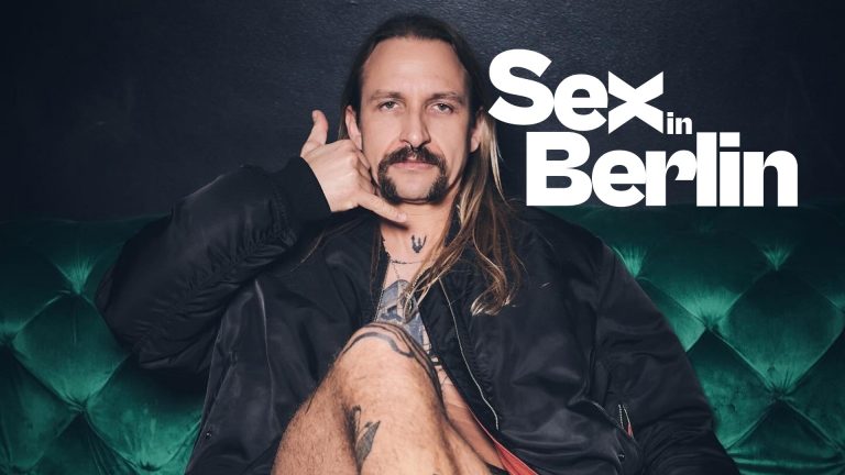 Mann mit langen Haaren und Schnurrbart sitzt auf einer grünen Couch, hält eine Hand an sein Ohr, Text "Sex in Berlin" im...