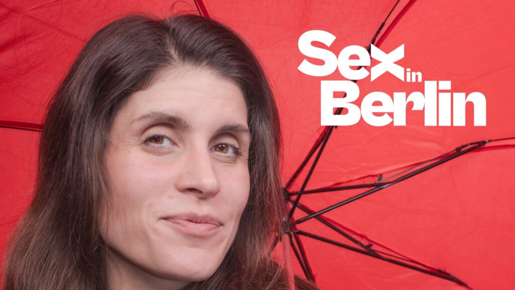 WEBSITE COVER - Sex in Berlin Frau mit langen, braunen Haaren lächelt vor rotem Hintergrund mit einem Schirm, Text "Sex in Berlin" daneben.