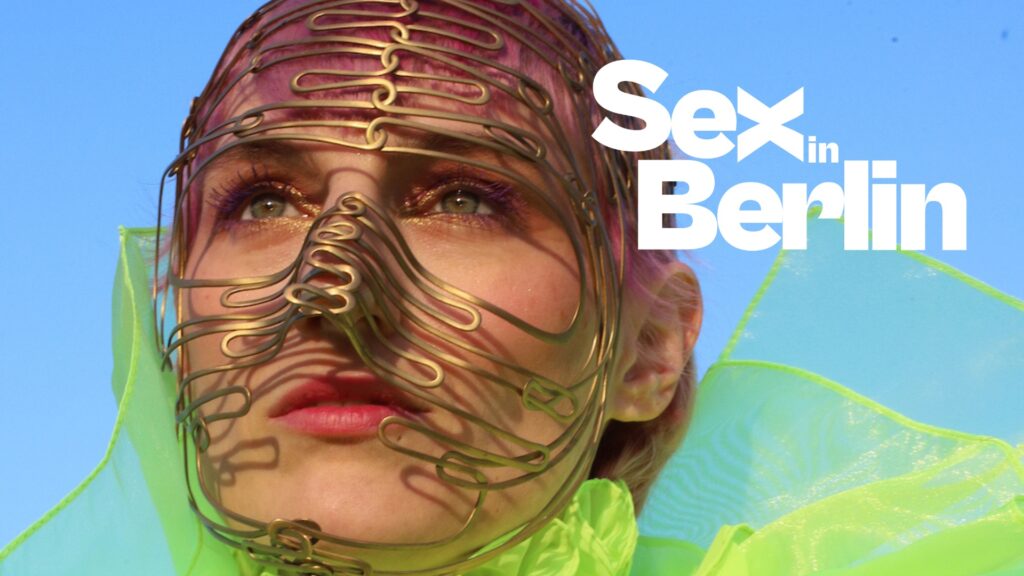 Close-up of a person wearing a metal wire mask against a bright blue sky, with bold white title 'Sex in Berlin' on the right.