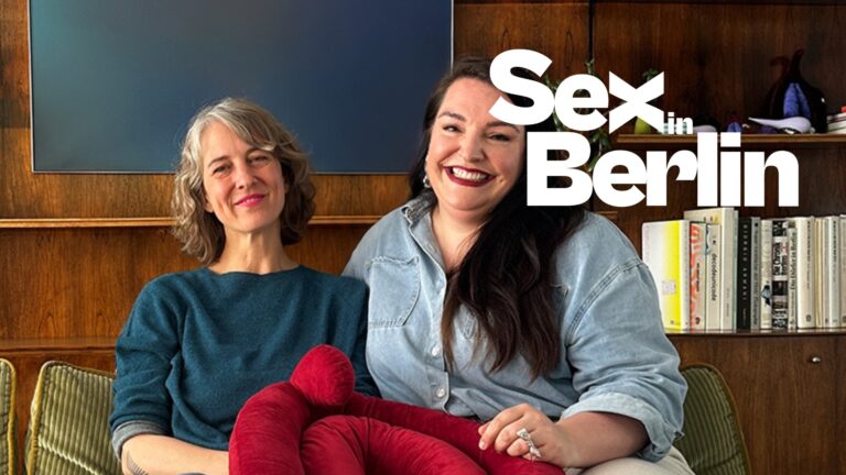 Two smiling women sit on a green sofa in a wood-paneled room, with a bold 'Sex in Berlin' overlay text.